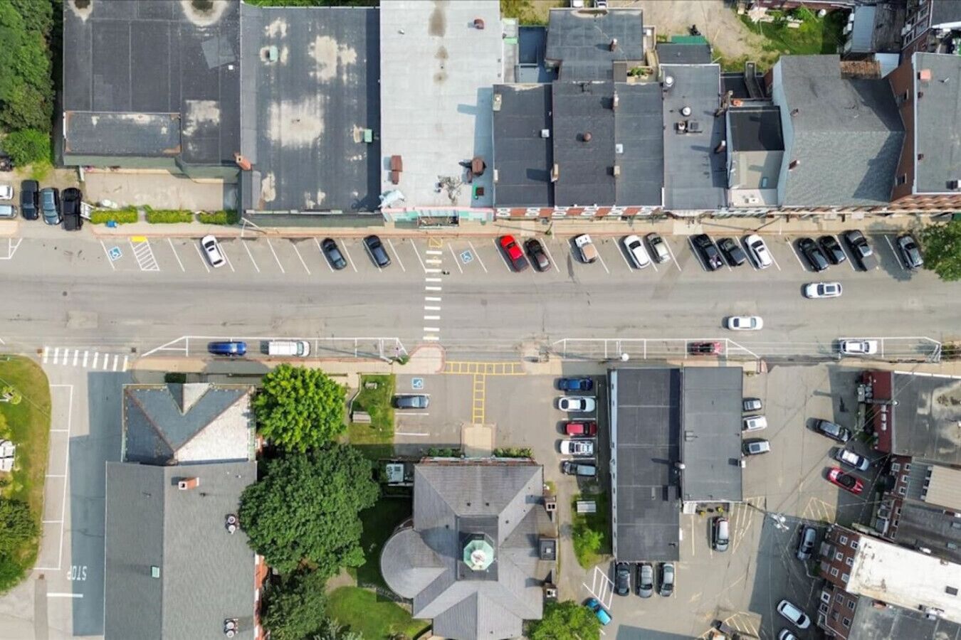 An aerial view of a parking lot with multiple rows of parked cars, a central pedestrian crossing, and surrounding buildings with various roof designs.