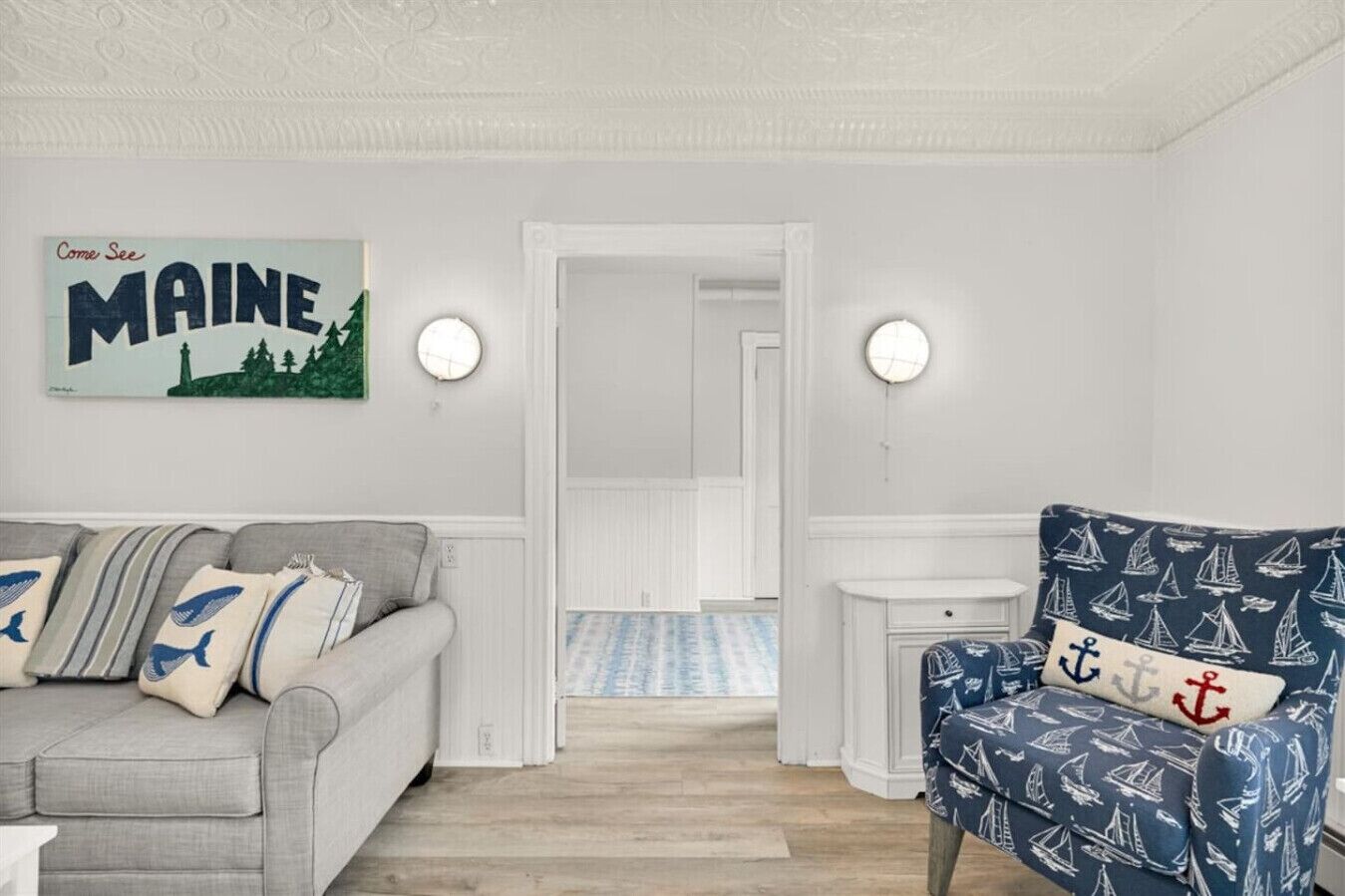 Une pièce avec un canapé gris, un fauteuil bleu orné de motifs marins, une pancarte indiquant « Venez découvrir le Maine », et un couloir menant à une autre pièce.