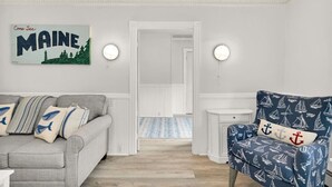 A room with a grey sofa, a blue armchair with nautical patterns, a 'Come See Maine' sign, and a hallway leading to another room. - Phoenix Nest - Downtown Apartment in Belfast (Belfast)