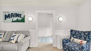 Une pièce avec un canapé gris, un fauteuil bleu orné de motifs marins, une pancarte indiquant « Venez découvrir le Maine », et un couloir menant à une autre pièce.