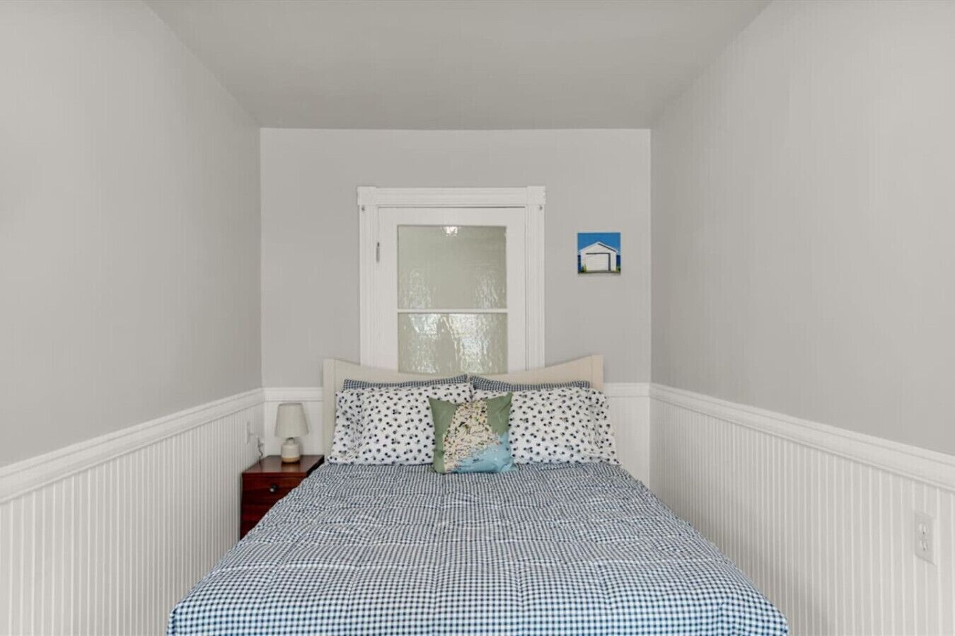 A bedroom with a checkered bedspread, a nightstand with a lamp, a window with a view, and a small picture on the wall.