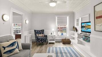 Un salon comprenant un canapé, un fauteuil, un téléviseur, une bibliothèque et une fenêtre avec des stores.