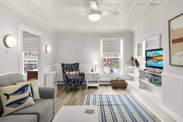 A living room with a sofa, armchair, TV, bookshelf, and a window with blinds.