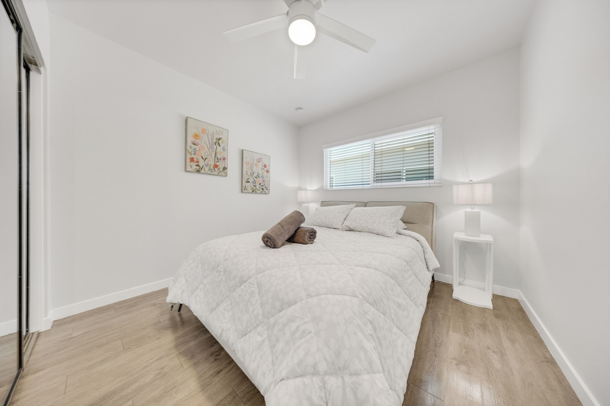 A bedroom with a bed, a ceiling fan, a window with blinds, and two wall-mounted lamps.