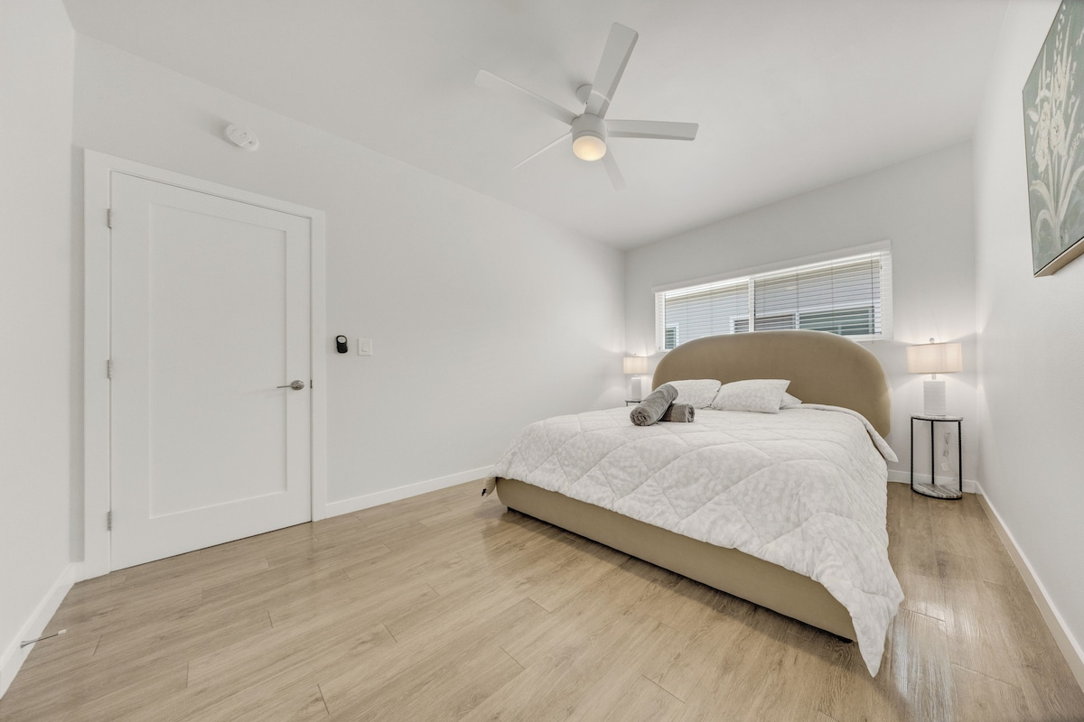 A bedroom with a bed, a ceiling fan, a window with blinds, and a nightstand with a lamp.