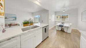 A modern kitchen with white cabinetry, a built-in dishwasher, and a dining area with a white table and chairs. - OneLuxStay Coastal 2BR Near Beach (Torrance)