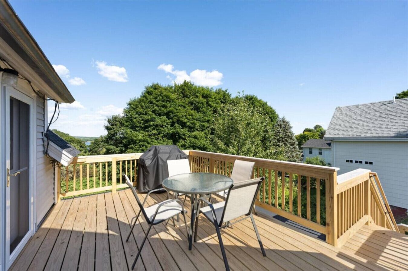 Une terrasse en bois avec une table et des chaises, un barbecue et une vue sur un arbre et une maison.