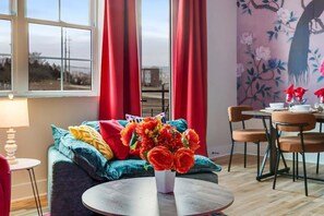 Ein Zimmer mit einem runden Tisch, einer Couch mit bunten Kissen, einer Blumenvase und einem Essbereich mit Stühlen und einem Tisch.