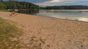 En strand med ett picknickbord, stilla vatten, båtar och en kustlinje som består av skog.