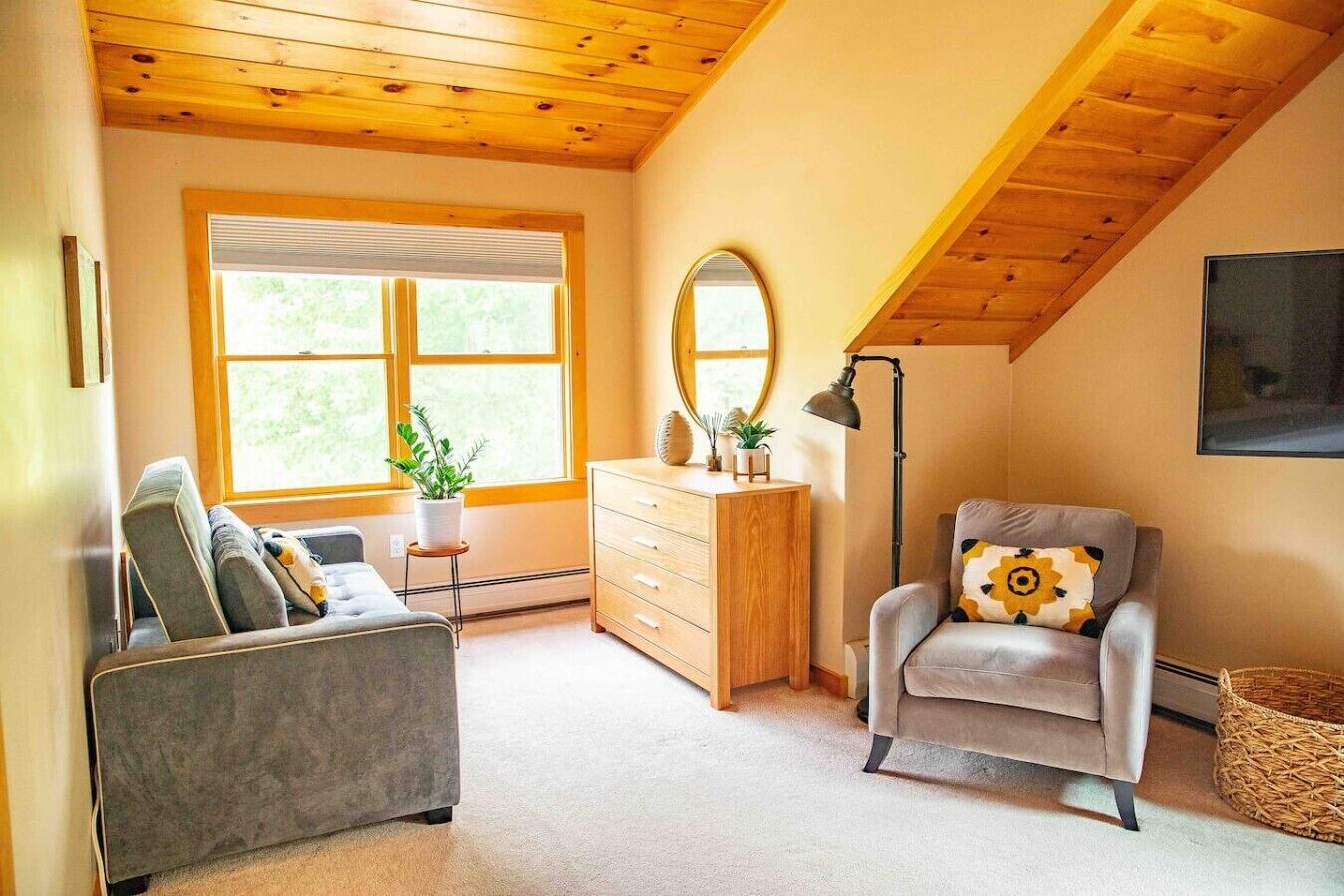Une pièce chaleureuse avec un plafond mansardé, un canapé, un fauteuil, une commode et une grande fenêtre donnant sur un paysage verdoyant.