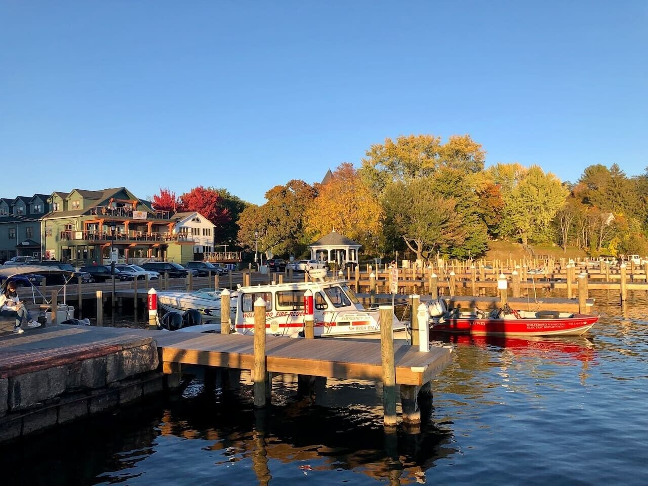 A waterfront marina with docked boats, a gazebo, and buildings in the background.