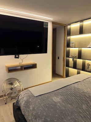 Ein modernes Schlafzimmer mit Bett, Wand-Fernseher, Nachttisch und integriertem Regal mit Beleuchtung.