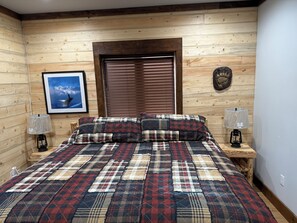 A wooden-paneled room with a bed, two bedside lamps, a framed picture, and a window with blinds. - SOLDOTNA SILVER TIP LODGE — Authentic Alaska Log Lodge Near World-Class Fishing (Soldotna)