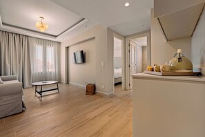 A modern hotel room with a sofa, a small table, a TV mounted on the wall, and a kitchenette area.