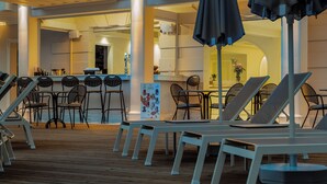 A sunlit outdoor seating area with lounge chairs and tables, a bar area in the background, and a wooden deck. - Royal Nidri Suites (Nidri)