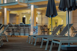 A sunlit outdoor seating area with lounge chairs and tables, a bar area in the background, and a wooden deck. - Royal Nidri Suites (Nidri)