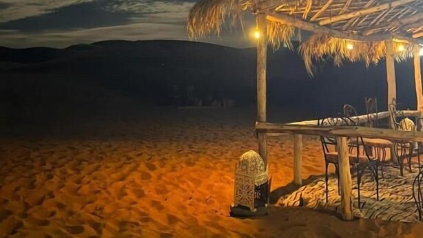 A beach scene with a thatched-roof structure, a table with chairs, and a lantern. - Desert Camel Trekking Camp (Taouz)