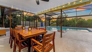 Un patio al aire libre con una mesa y sillas de madera, un ventilador de techo, luces colgantes y vista a una piscina.
