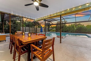 An outdoor patio with a wooden table and chairs, a ceiling fan, string lights, and a view of a swimming pool. - Free Heated Pool & Spa : Game Room: 5mi to beaches: Lakefront: IMG: AMI (Bradenton)