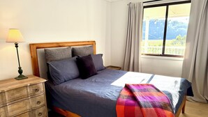 Ein Bett mit hölzernem Gestell, grauer Bettwäsche und einer bunten Decke.