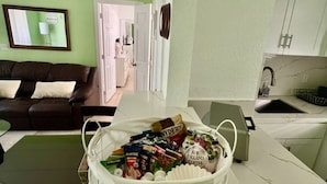A living room with a brown leather sofa, a white coffee table with a basket of snacks, a kitchen area with a sink and cabinets, and a hallway leading to a door.