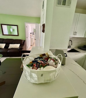 Una sala de estar con un sofá de cuero marrón, una mesa de centro blanca con una cesta de aperitivos, una zona de cocina con fregadero y armarios, y un pasillo que lleva a una puerta.