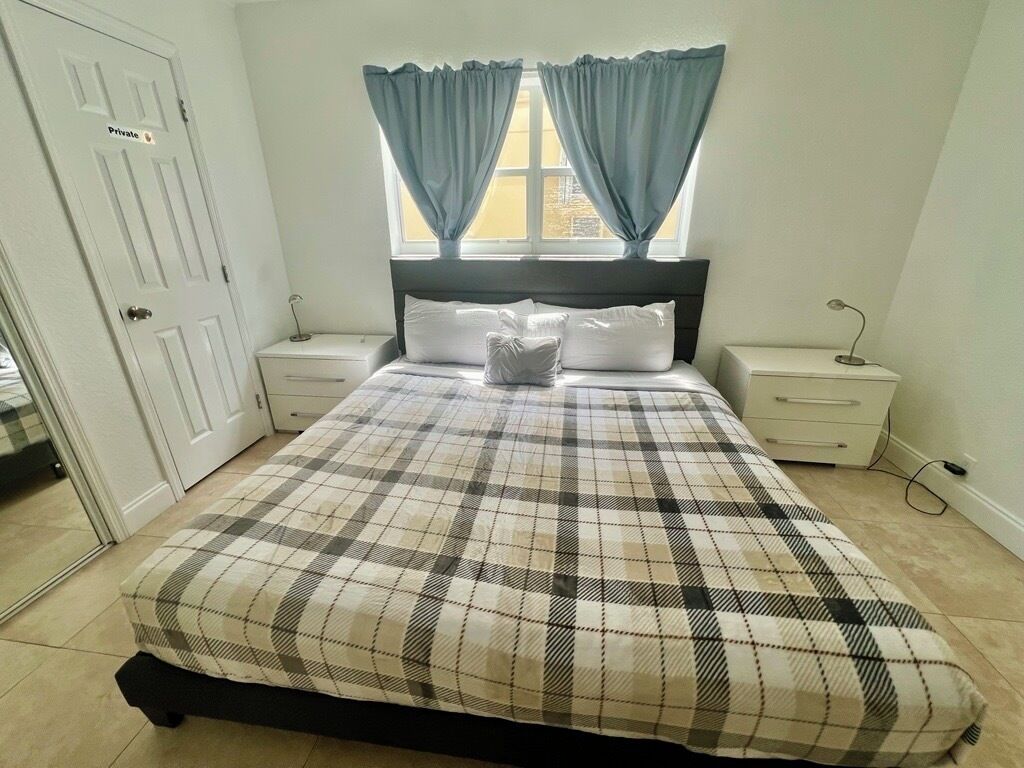 Un dormitorio con cama, dos mesitas de noche, una mesilla auxiliar, una ventana con cortinas y una puerta con la leyenda 'Privado'.
