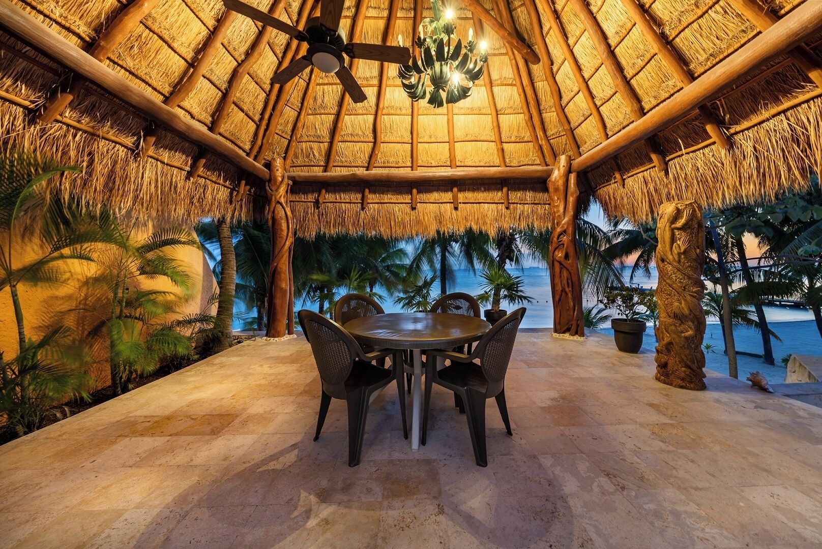 A thatched-roof outdoor dining area with a table and chairs, surrounded by palm trees and a view of the ocean.