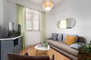 Ein modernes Wohnzimmer mit Flachbildfernseher, einer Couch mit Kissen, einem runden Couchtisch und einem Spiegel an der Wand.