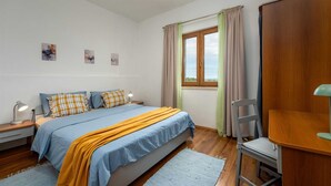 Ein Schlafzimmer mit einem Bett, Nachttischen, einem Sessel, einem Schreibtisch, einem Fenster mit Vorhängen und Wandbildern.