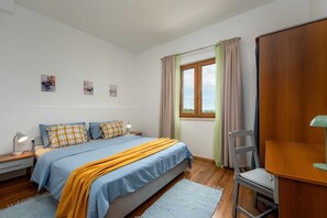 Ein Schlafzimmer mit einem Bett, Nachttischen, einem Sessel, einem Schreibtisch, einem Fenster mit Vorhängen und Wandbildern.