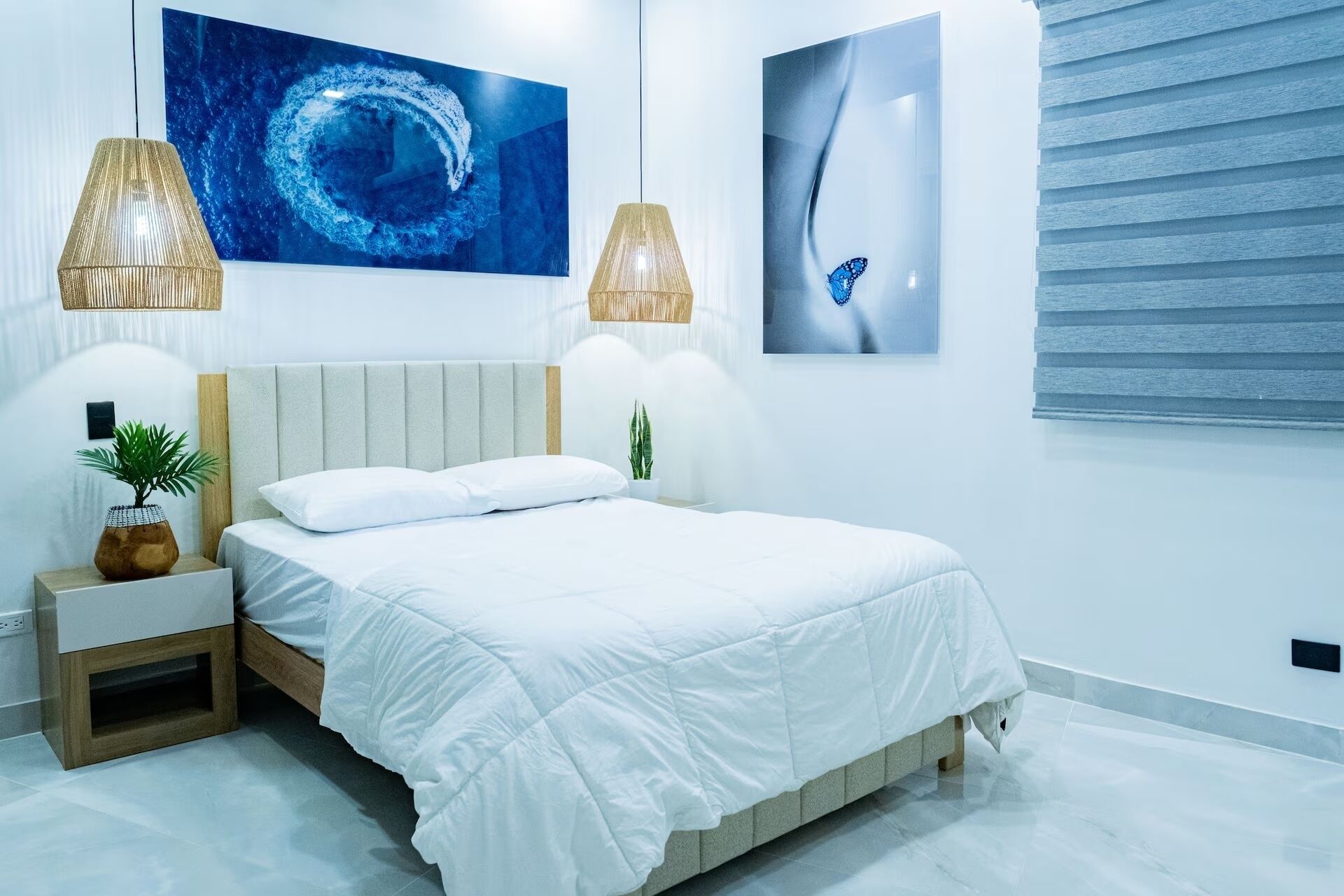 A modern bedroom with a bed, two wall-mounted lamps, a potted plant, and two framed artworks.