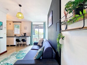 A modern living room with a sofa, a dining area with chairs, a kitchen, and a large window. - Natur’Azur - AC - Sainte-Clotilde -Patio (Saint-Denis)