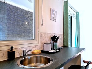 A kitchen with a stainless steel sink, a window with blinds, a marble backsplash, and a wall-mounted spice rack. - Natur’Azur - AC - Sainte-Clotilde -Patio (Saint-Denis)