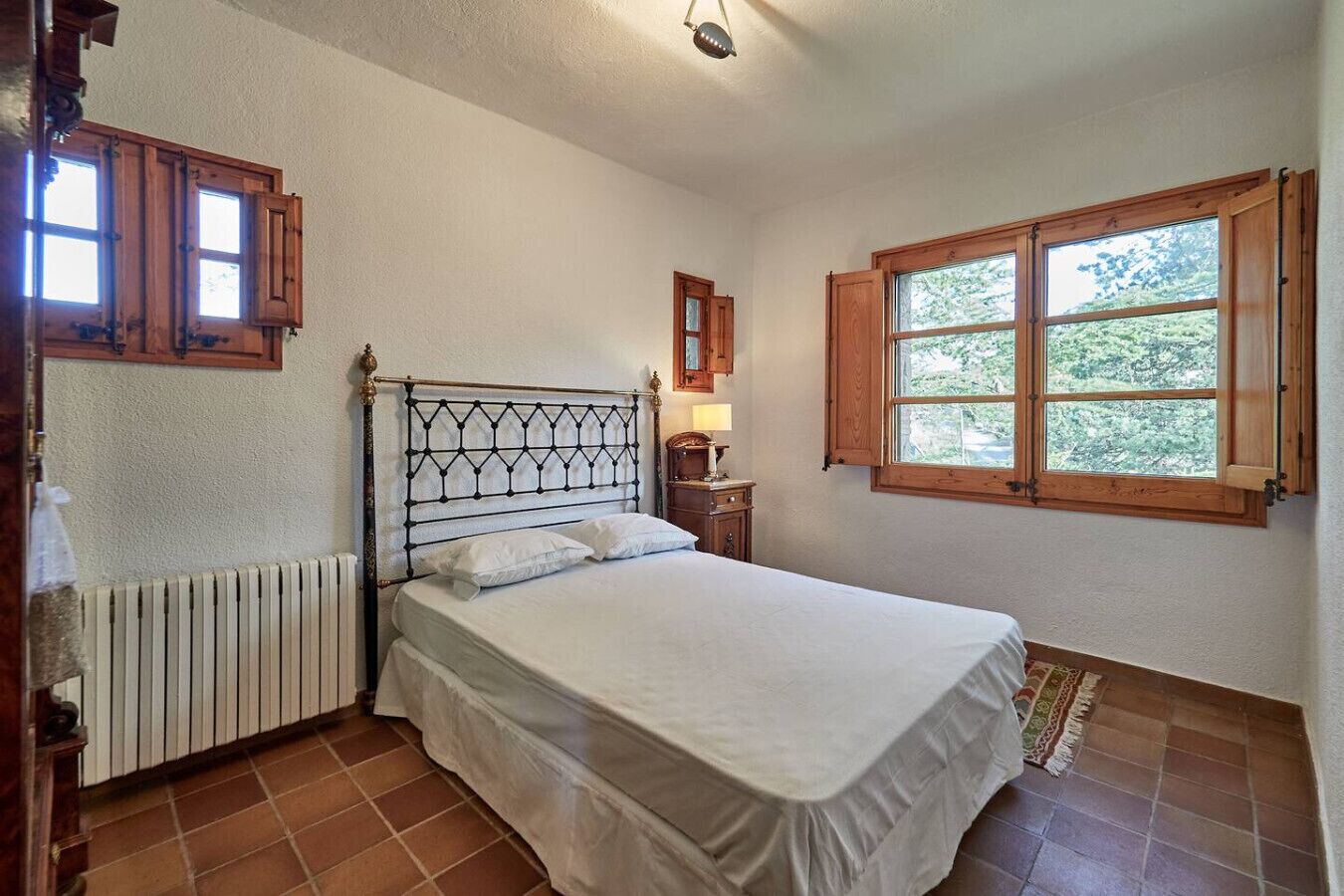 Ein Schlafzimmer mit Bett, Holzschränken, einem Heizkörper, einem Fenster mit Blick auf Bäume und einem kleinen Nachttisch mit Lampe.