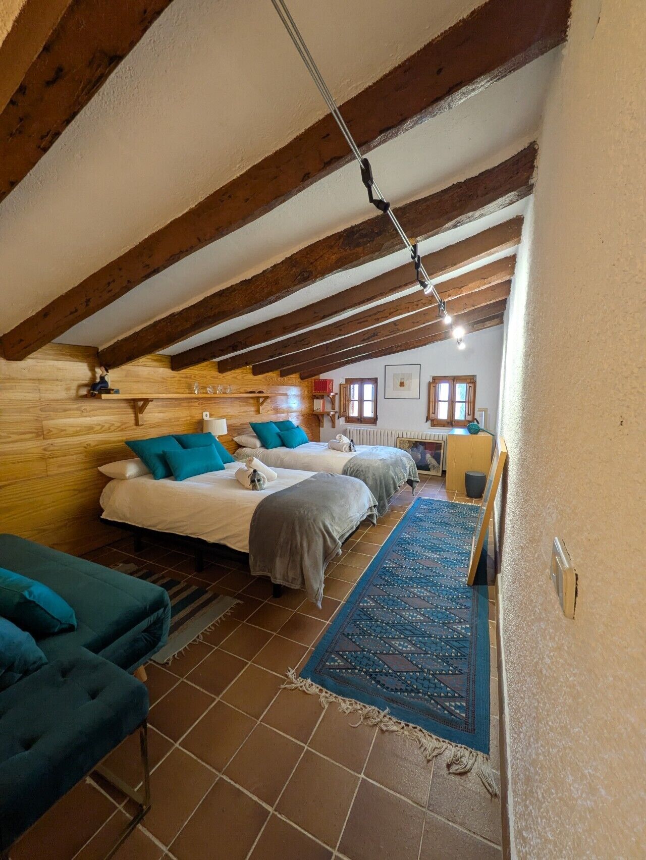 Quarto com duas camas, um sofá, tapete estampado, vigas de madeira e uma janela.