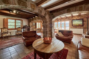 A rustic interior with stone walls, wooden beams, leather sofas, and a round wooden table.