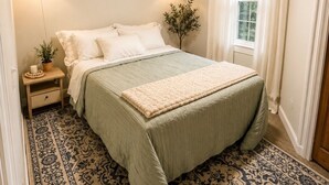 Un dormitorio con una cama, un ventilador de techo, una ventana con cortinas, una mesita de noche con lámpara y un cuadro colgado en la pared.