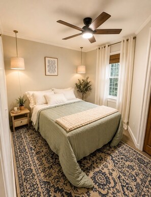 A bedroom with a bed, a ceiling fan, a window with curtains, a bedside table with a lamp, and a wall-mounted picture.