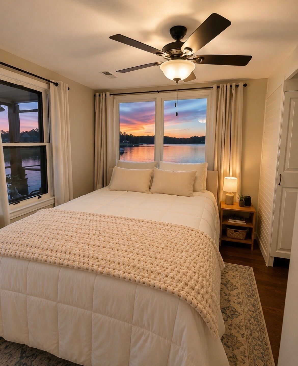 Une chambre à coucher avec un lit, un ventilateur de plafond, une table de chevet avec une lampe, et une vue sur un lac à travers deux fenêtres.