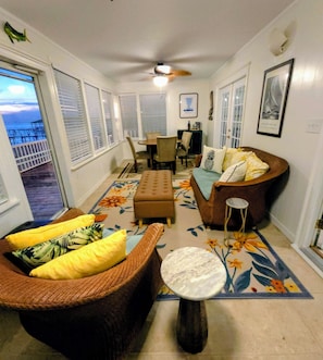 Ein Wohnzimmer mit einer Couch, Hocker und einem kleinen runden Tisch. Es gibt eine Essecke mit Tisch und Stühlen. Durch große Fenster hat man einen Ausblick.
