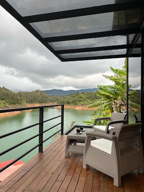 A wooden deck with wicker furniture overlooking a calm body of water and a forested hillside. - Boreal Glamping (Guatape)