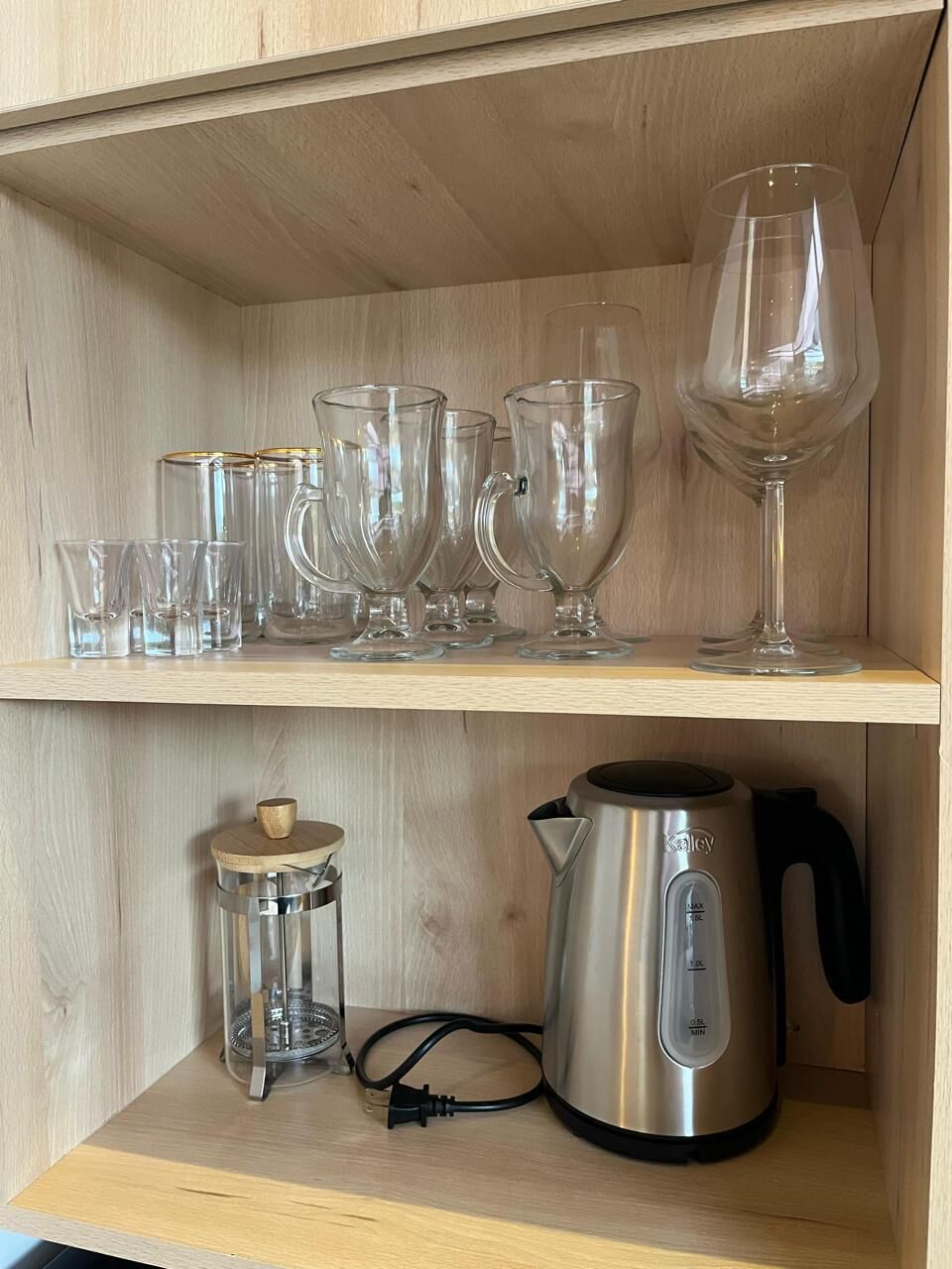 Una estantería de madera con diferentes vasos, una jarra de vidrio, una tetera de vidrio y una hervidora.