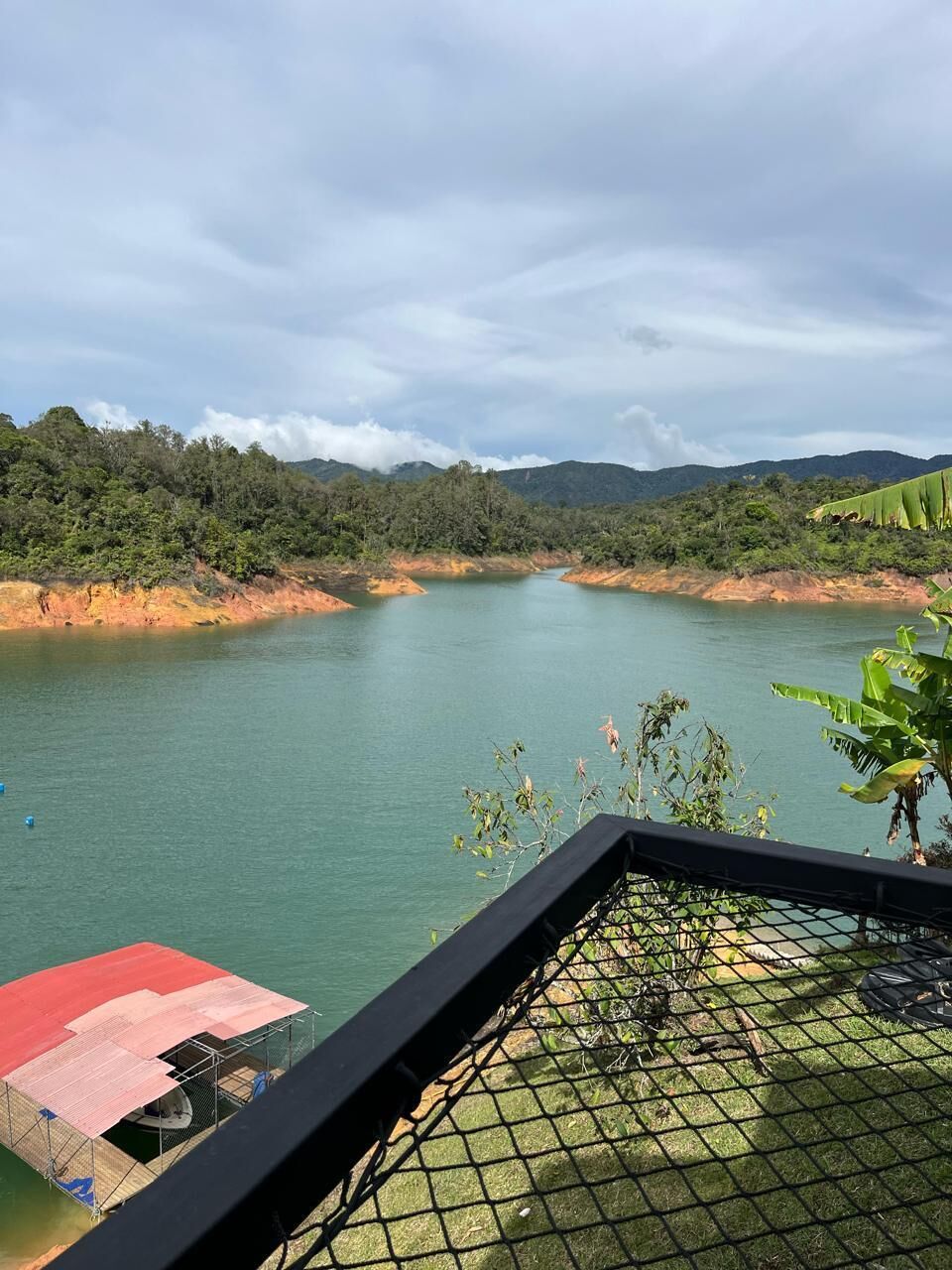 A view of a calm lake with a red-roofed structure on the left, surrounded by greenery and hills.