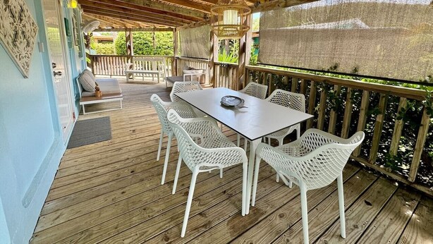 Um deck de madeira ao ar livre com mesa e cadeiras, uma área coberta com teto e vista para o verde.