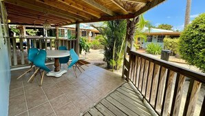 Uma área externa coberta com um deck de madeira, uma mesa redonda com cadeiras e vista para edifícios e vegetação.