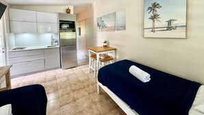 Quarto de hotel compacto com cozinha compacta, uma cama com toalha enrolada, uma mesa pequena com cadeiras e uma pintura com tema de praia na parede.