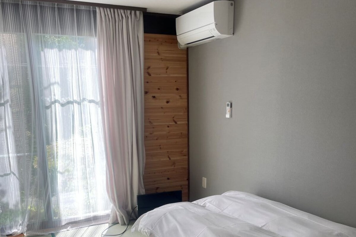 A room with a bed, a window with sheer curtains, and an air conditioning unit on the wall.