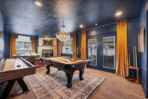 A room with a pool table, a billiards table, a chandelier, and a fireplace. - Game Rooms, Hot Tub & Fire Pit (Arvada)