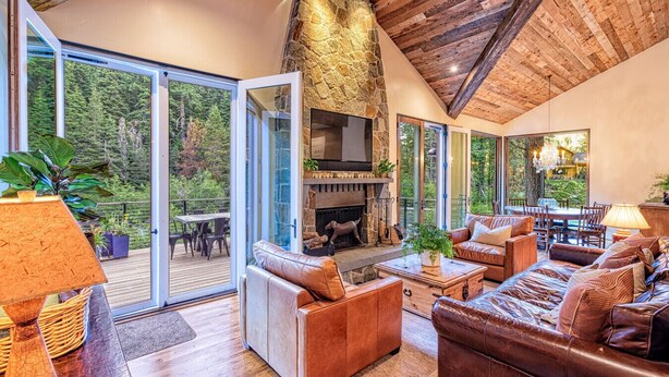 A spacious living area with a stone fireplace, leather sofas, a dining table, and a view of a forested area through large glass doors.